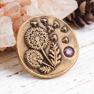 Peut inclure: Un bouton rond en métal couleur laiton antique présentant un motif en relief de deux têtes de pissenlit avec des graines flottantes et des feuilles. Une petite pierre précieuse rose foncé est incrustée à la surface du bouton.
