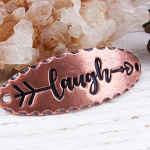 May include: Copper oval charm with an arrow pointing to the right and the word "laugh" written in cursive.