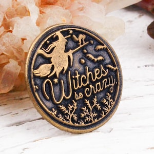 May include: A round, dark-toned metal pin with a raised gold-coloured relief design. The pin features a silhouette of a witch flying on a broomstick, stars, and the text Witches be crazy in a stylised script font, accented by small leafy branches.