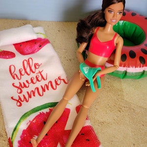 May include: A doll wearing a red swimsuit and a green visor is laying on a watermelon-patterned beach towel. The towel says "hello sweet summer".