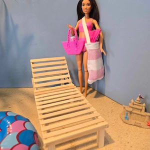 May include: A pink and white doll with long black hair is standing next to a wooden lounge chair. The doll is holding a pink beach bag and a pink towel. The doll is standing on a sandy surface with a sandcastle in the background.