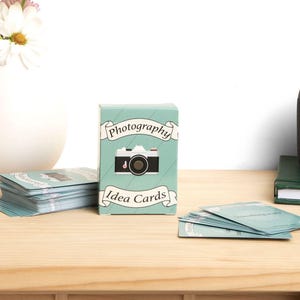 May include: A teal blue box with a vintage camera illustration and the text "Photography Idea Cards". The box is sitting on a wooden surface with a stack of cards next to it.