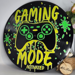Może przedstawiać: Czarna grafika na płycie winylowej z neonowo-zielonym i żółtym wzorem kontrolera do gier. Na płycie namalowano napisy „GAMING MODE ACTIVATED”. Zielone plamy farby dodają dekoracyjnego charakteru. Unikalny element dla graczy.