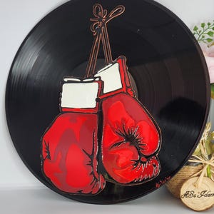 Puede incluir: Un disco de vinilo negro con un par de guantes de boxeo rojos con puños blancos. Los guantes están suspendidos por cuerdas marrones. La obra de arte es una pieza audaz y llamativa, perfecta para los entusiastas del deporte.