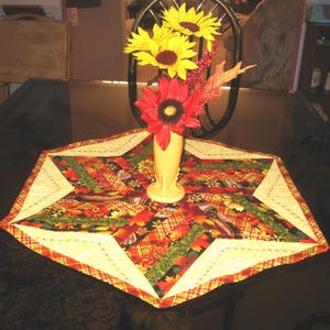 May include: A decorative table centerpiece featuring a star-shaped quilted table runner with a floral and plaid pattern in autumn colors. A yellow vase holds red and yellow sunflowers and fall foliage.