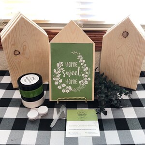 May include: A craft kit for making a home sweet home sign. The kit includes three unfinished wooden houses, a green sign with the text "Home Sweet Home" in white, a jar of black paint, a jar of white paint, a jar of green paint, a small spatula, and a business card.