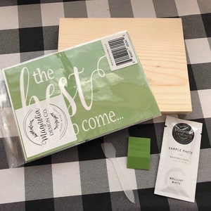 May include: A green stencil with the text "the best is to come..." and a white circle with the text "Magnolia Design Co."  A wooden sign, a green sample of paint, and a white packet of sample paste are also included.