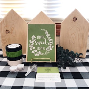 May include: A craft kit for making a home sweet home sign. The kit includes three unfinished wooden houses, a green stencil with the text "Home Sweet Home" in a white wreath, black and green paint, a paint scraper, and a business card.