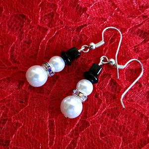 May include: A pair of silver snowman earrings with white pearl beads and black top hats. The earrings are on a red lace background.
