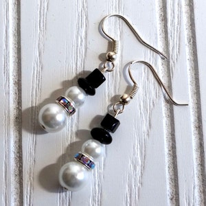 May include: A pair of silver snowman earrings with black beads and white pearl beads. Each snowman has a black bead for a hat and a rhinestone for a button.