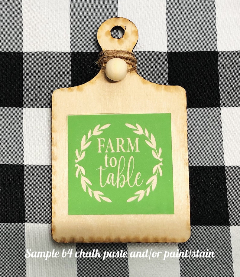 May include: A wooden cutting board with a green square in the center. The square has a white wreath with the words "FARM to table" in the center.