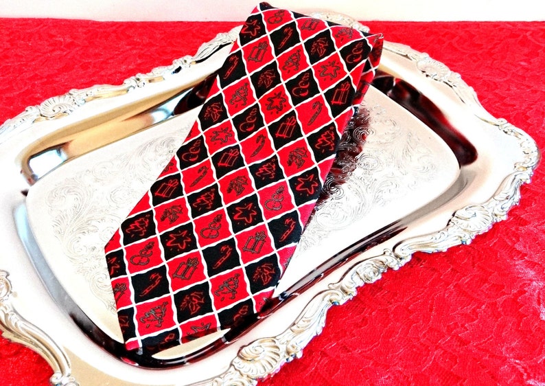 May include: A red and black patterned tie with a Christmas theme. The tie features a repeating pattern of Christmas trees, stars, presents, candy canes, and sleighs. The tie is lying on a silver serving tray.