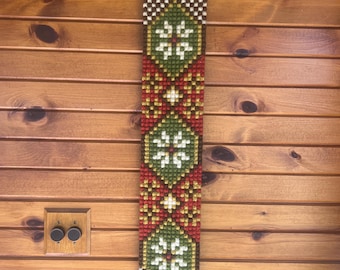 May include: A long, vertical needlepoint wall hanging featuring a repeating geometric pattern of green diamonds, white star-like motifs, and red and brown accents. The textile is suspended from a decorative brass hanger at the top and bottom.