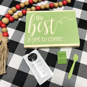May include: A green wooden sign with the text "the best is yet to come..." in white lettering. The sign is sitting on a black and white checkered surface. There is a small white packet of sample paste and a green spatula on the surface.