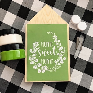 May include: A wooden house-shaped sign with a green background and white lettering that reads "Home Sweet Home" surrounded by a wreath of white leaves. The sign is on a black and white checkered surface with paint jars and a paint scraper.