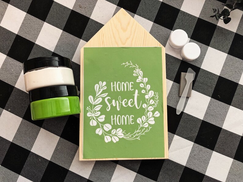 May include: A wooden house-shaped sign with a green background and white lettering that reads "Home Sweet Home" surrounded by a wreath of white leaves. The sign is on a black and white checkered surface with paint jars and a paint scraper.