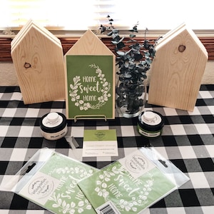 May include: A set of three unfinished wooden house-shaped signs with a green and white stencil that reads "Home Sweet Home" and a wreath of leaves. The stencil is on a green background and is surrounded by a gold frame. The signs are on a black and white checkered tablecloth with paint jars and a vase of eucalyptus.