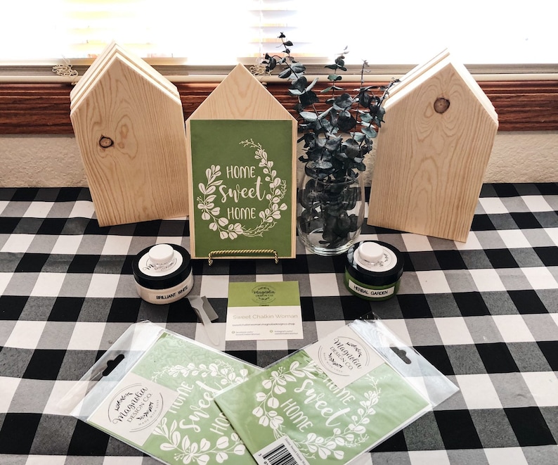 May include: A set of three unfinished wooden house-shaped signs with a green and white stencil that reads "Home Sweet Home" and a wreath of leaves. The stencil is on a green background and is surrounded by a gold frame. The signs are on a black and white checkered tablecloth with paint jars and a vase of eucalyptus.
