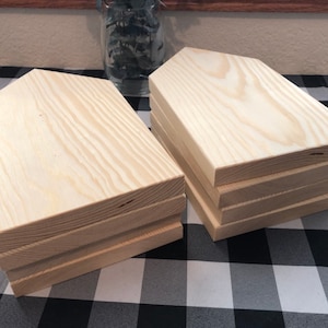 May include: A stack of six unfinished wooden house-shaped signs. The signs are made of light-colored wood and have a slightly rough texture. They are stacked on top of each other, with the top sign being the largest and the bottom sign being the smallest.