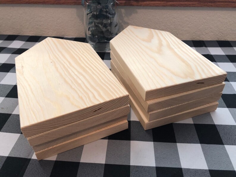 May include: A stack of six unfinished wooden house-shaped signs. The signs are made of light-colored wood and have a slightly rough texture. They are stacked on top of each other, with the top sign being the largest and the bottom sign being the smallest.