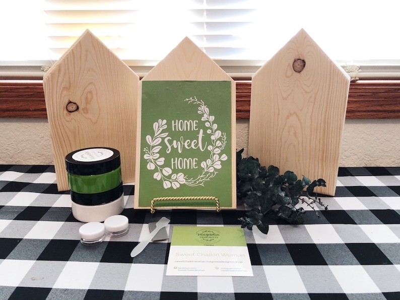 May include: A craft kit for making a home sweet home sign. The kit includes three unfinished wooden houses, a green stencil with the text "Home Sweet Home" in a white wreath, black and green paint, a paint scraper, and a business card.