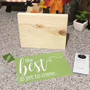 May include: A green stencil with the text "the best is yet to come..." in white lettering. The stencil is on a wooden surface with a small wooden sign and a small white pot with a green plant.