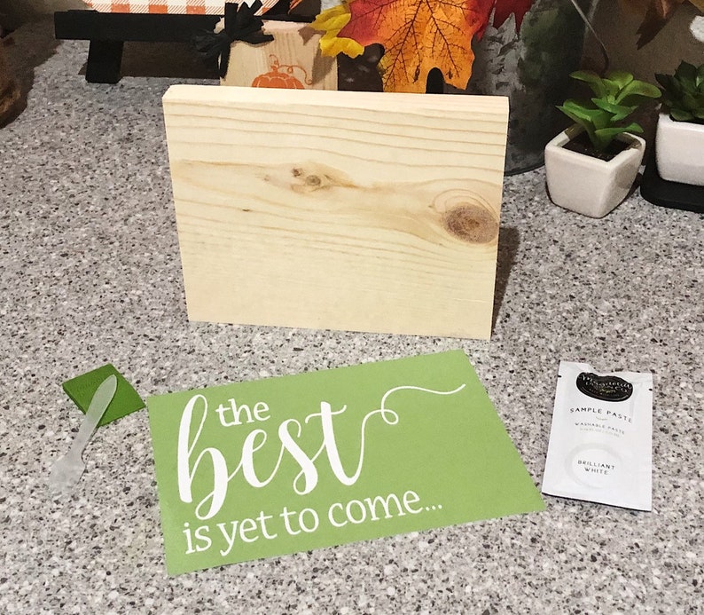 May include: A green stencil with the text "the best is yet to come..." in white lettering. The stencil is on a wooden surface with a small wooden sign and a small white pot with a green plant.