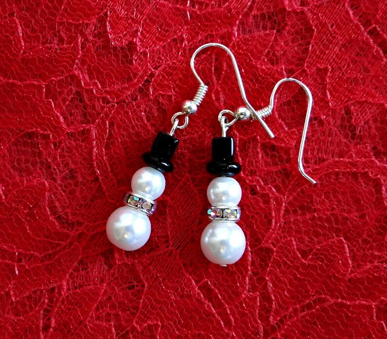 May include: A pair of silver snowman earrings with black hats and white pearl beads. The earrings have a silver hook fastening.