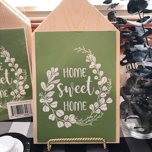 May include: A wooden house-shaped sign with a green background and white lettering that reads "Home Sweet Home". The sign is decorated with a white floral wreath. The sign is on a gold stand and is sitting on a black and white checkered surface.