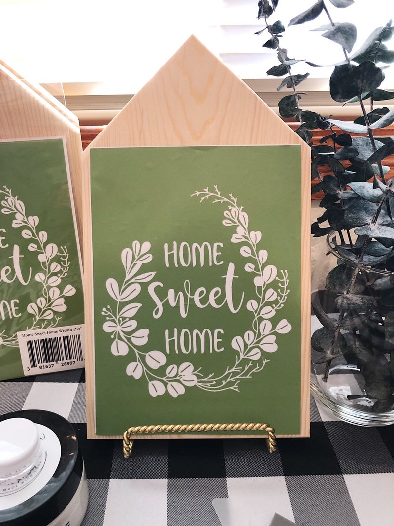 May include: A wooden house-shaped sign with a green background and white lettering that reads "Home Sweet Home". The sign is decorated with a white floral wreath. The sign is on a gold stand and is sitting on a black and white checkered surface.