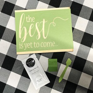 May include: A green wooden sign with the text "the best is yet to come..." in white lettering. A small white packet of sample paste and a green spatula are also pictured.