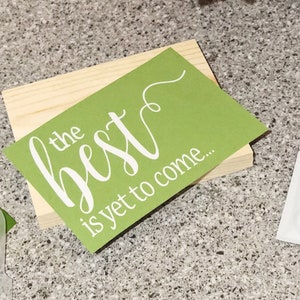 May include: A wooden sign with a green background and white lettering that reads "the best is yet to come..." The sign is sitting on a table with a small container of white paint and a green paint scraper.