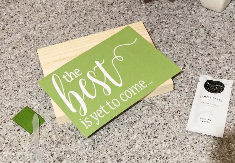 May include: A wooden sign with a green background and white lettering that reads "the best is yet to come..." The sign is sitting on a table with a small container of white paint and a green paint scraper.