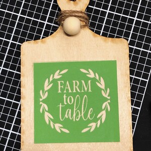 May include: A wooden cutting board with a green square sign that says "FARM to table" in white lettering. The sign is surrounded by a white wreath of leaves. The cutting board has a wooden handle with a small wooden knob at the top. The cutting board is on a black and white grid background. The word "Sample" is written in white on the bottom right corner of the cutting board.