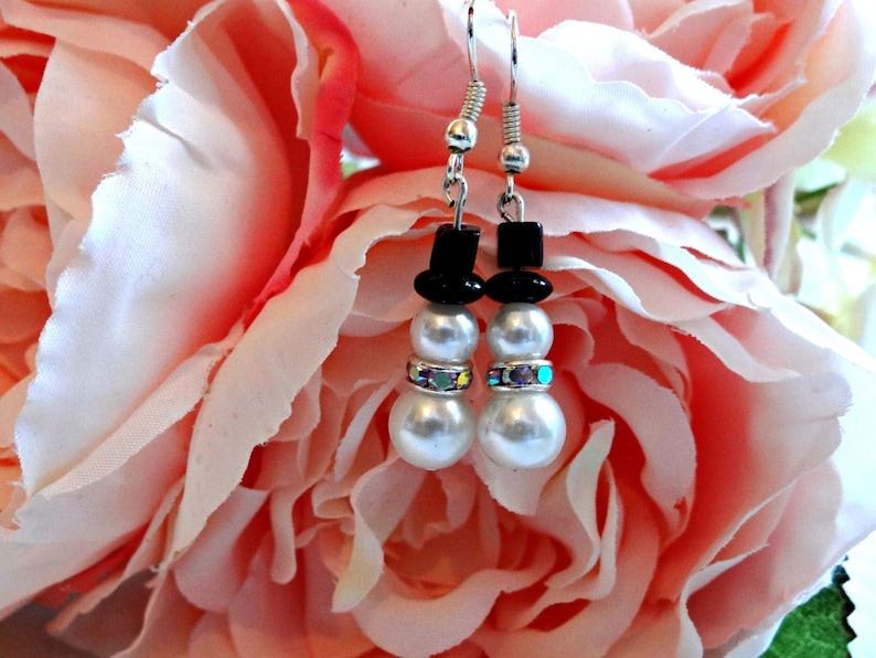 May include: A pair of silver snowman earrings with black hats and white pearl beads. The earrings are accented with iridescent rhinestones.