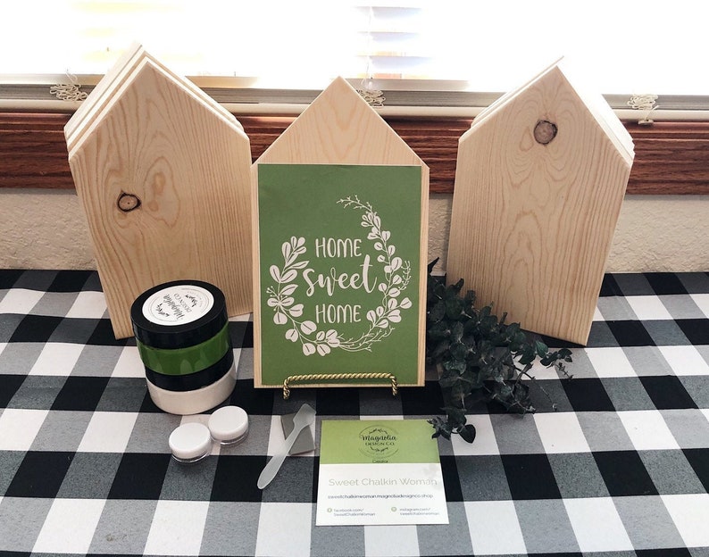 May include: A craft kit for making a home sweet home sign. The kit includes three unfinished wooden houses, a green sign with the text "Home Sweet Home" in white, a jar of black paint, a jar of white paint, a jar of green paint, a small spatula, and a business card.