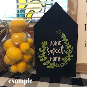 May include: Black wooden house-shaped sign with a white inscription that reads "Home Sweet Home" surrounded by a green leafy wreath.
