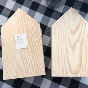 May include: Two unfinished wooden house-shaped signs. The sign on the left has a sticky note with the text "9 1/4"T 5 1/2"W 3/4" thick".