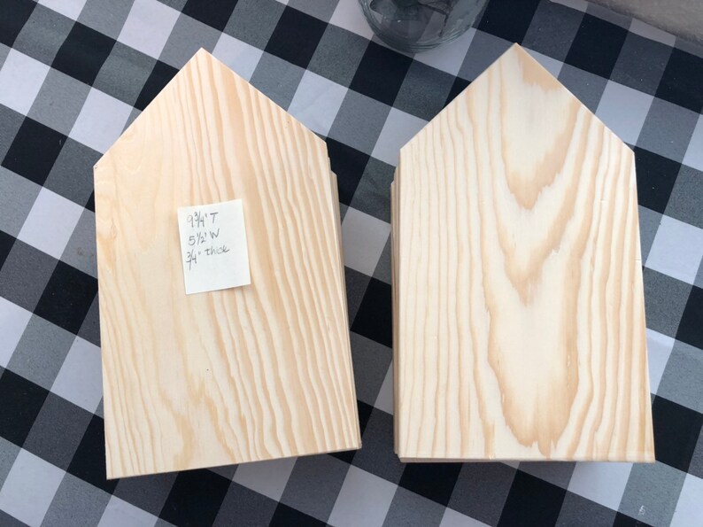 May include: Two unfinished wooden house-shaped signs. The sign on the left has a sticky note with the text "9 1/4"T 5 1/2"W 3/4" thick".