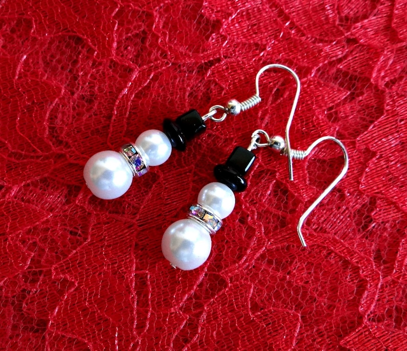 May include: A pair of silver snowman earrings with white pearl beads and black top hats. The earrings are on a red lace background.