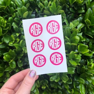 May include: A sheet of six hot pink monogram stickers with the letters "RGE" in a white, circular design. The stickers are held against a backdrop of green foliage.