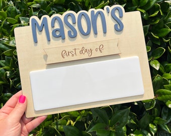 May include: A wooden sign with the name "Mason's" in blue letters and a banner that says "first day of". The sign has a white space for writing the date.