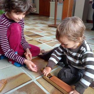 May include: Two children playing with wooden toys on a tiled floor. One child wears a striped maroon and white shirt and maroon pants. The other child wears a brown and white striped shirt and brown pants. The toys appear to be wooden blocks.