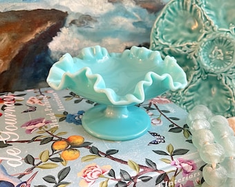 Op de afbeelding: Een turquoise blauwe glazen snoepschotel met een gekrulde rand en een voet. De schotel staat op een boek met een bloemenmotief.