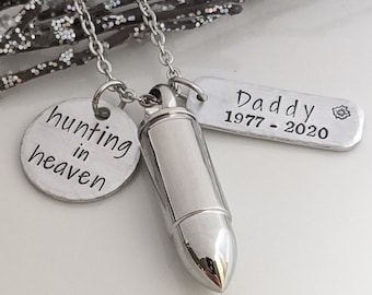 May include: A silver-toned necklace with three pendants. One pendant is a bullet-shaped urn, another is a round charm with the words "hunting in heaven", and the third is a rectangular tag with "Daddy 1977-2020".