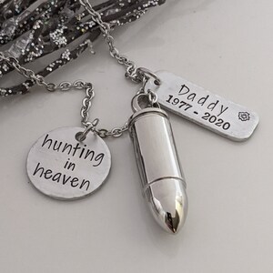 May include: Silver-toned memorial necklace with a bullet-shaped pendant, a round charm with "hunting in heaven" text, and a rectangular tag with "Daddy 1977-2020" engraved. The necklace is displayed on a white surface with glittery branches.