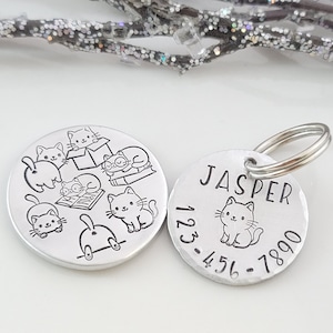 May include: Two silver pet ID tags with a cat theme. One tag has a design of multiple cats with a book and the text "123-456-7890". The other tag has the name "JASPER" and a single cat design.
