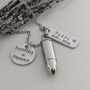 May include: A silver-colored necklace with three pendants. One pendant is a bullet-shaped urn, another is a round disc with the words "hunting in heaven", and the third is a rectangular tag with the word "Daddy" and the years "1977-2020".