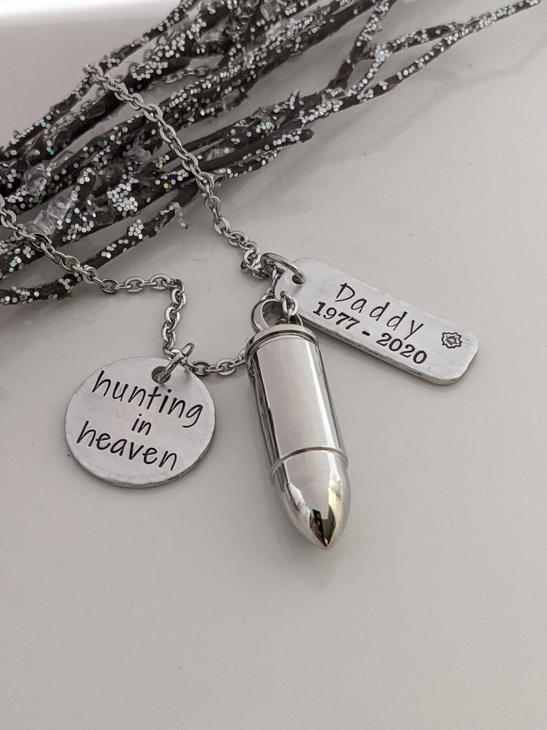 May include: A silver-colored necklace with three pendants. One pendant is a bullet-shaped urn, another is a round disc with the words "hunting in heaven", and the third is a rectangular tag with the word "Daddy" and the years "1977-2020".