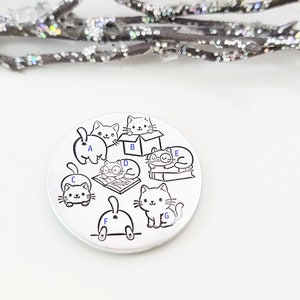 May include: A silver charm with a black and white illustration of seven cats in various poses. The cats are labeled with letters A through G.